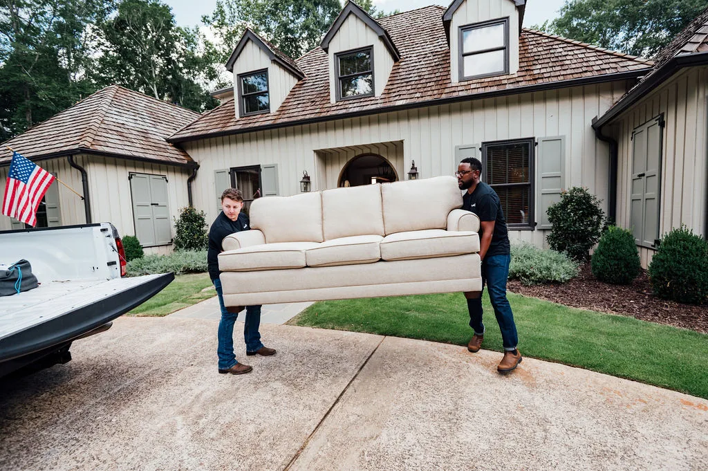 Two LoadUp Loaders carrying a cream three-seat couch across a driveway to load it onto a pickup truck