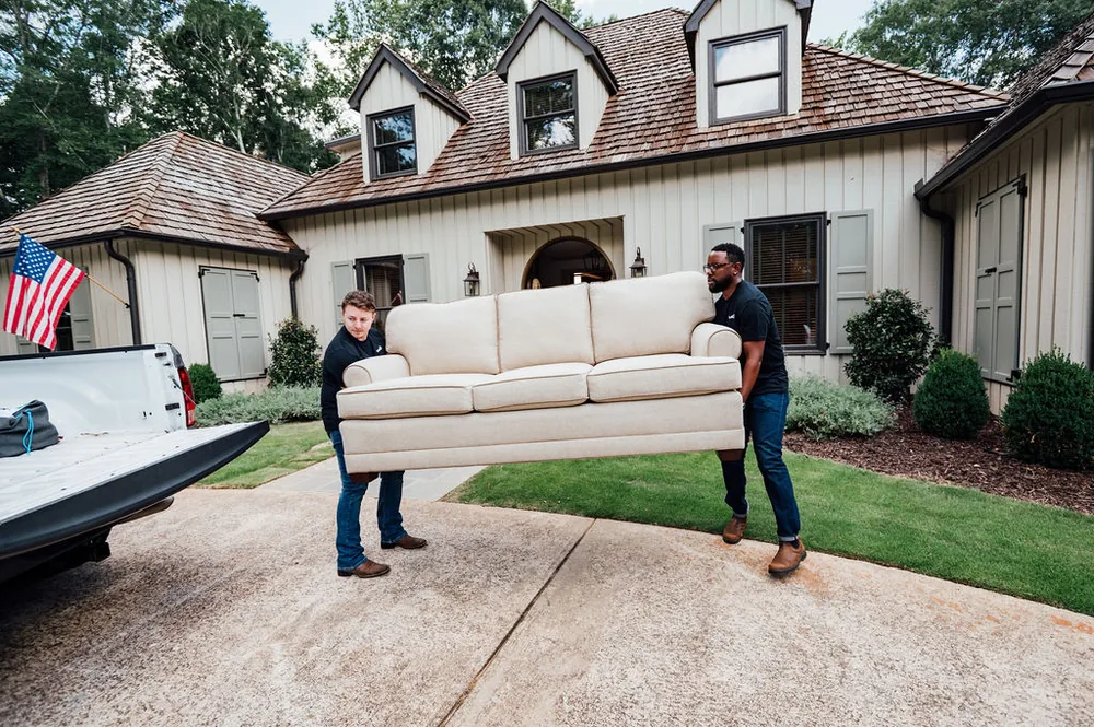 Two Furniture Disposal Plus Loaders carrying a large three-seat couch out through a doorway